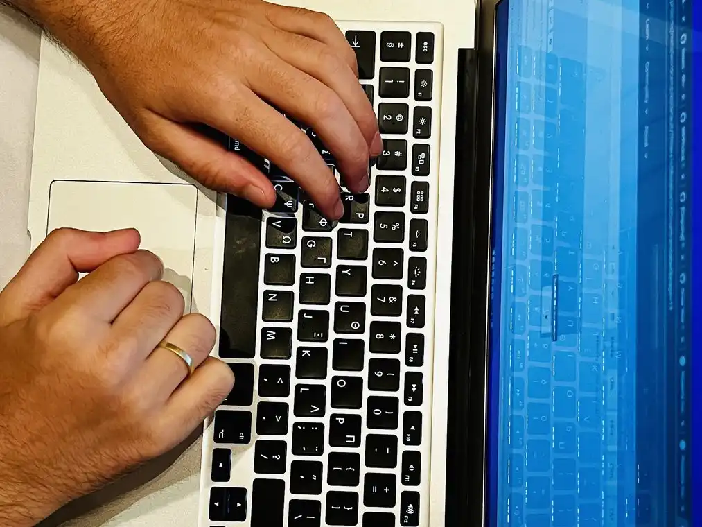 Two hands typing on a silver laptop, with a black phone beside it on a white desk