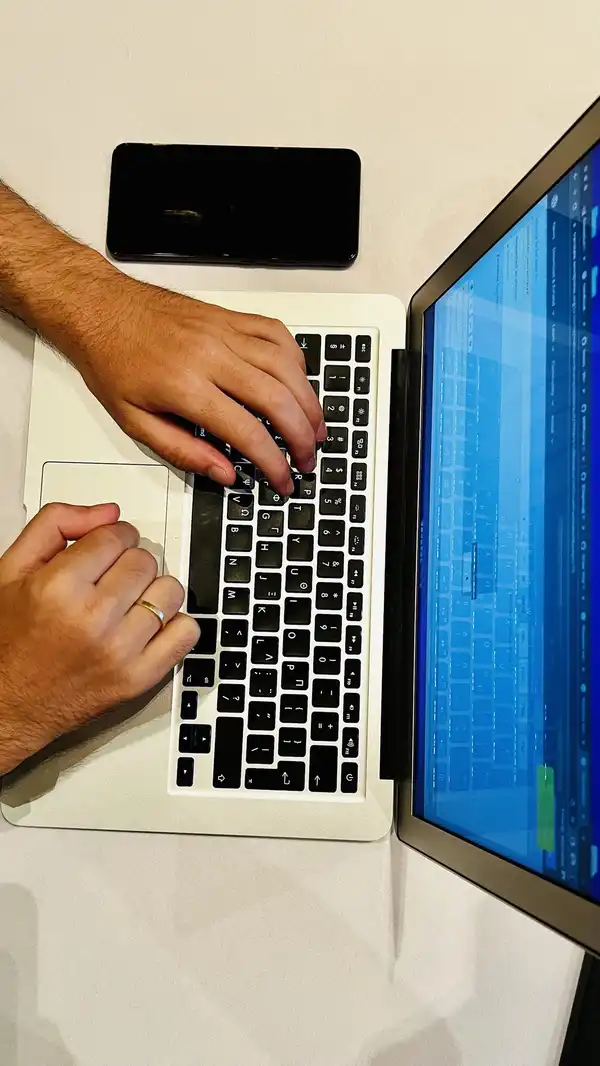Two hands typing on a silver laptop, with a black phone beside it on a white desk